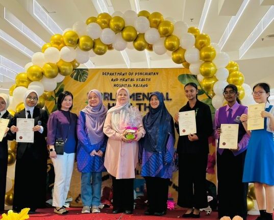 A group of individuals at a psychiatry and mental health event at Hospital Kajang, standing under a balloon arch and holding certificates.