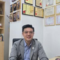 Dr. Henry Tan sitting at his office desk, wearing a gray blazer and glasses, with framed certificates and awards on the wall behind him.