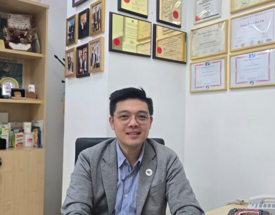 Dr. Henry Tan sitting at his office desk, wearing a gray blazer and glasses, with framed certificates and awards on the wall behind him.