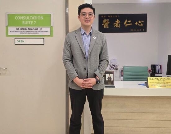 Dr. Henry Tan Chor Lip standing in front of his consultation room, dressed in professional attire, with a welcoming smile.