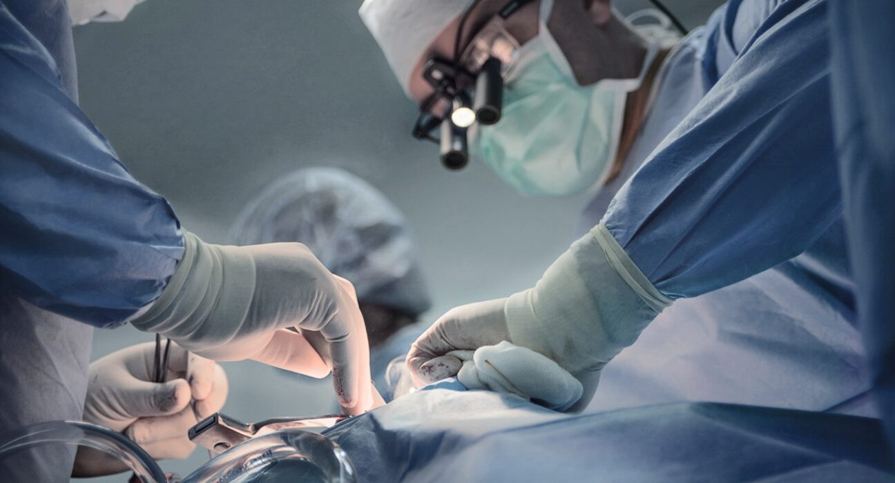 Coronary Artery Disease Singapore - A team of surgeons performing a surgical procedure in an operating room. The lead surgeon wears magnifying surgical loupes and a face mask, while assistants in sterile gloves carefully handle medical instruments.