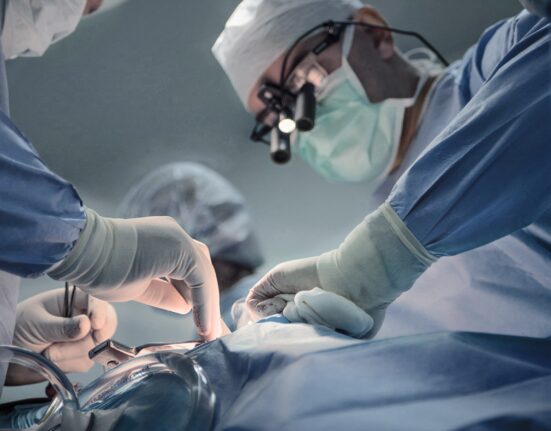Coronary Artery Disease Singapore - A team of surgeons performing a surgical procedure in an operating room. The lead surgeon wears magnifying surgical loupes and a face mask, while assistants in sterile gloves carefully handle medical instruments.