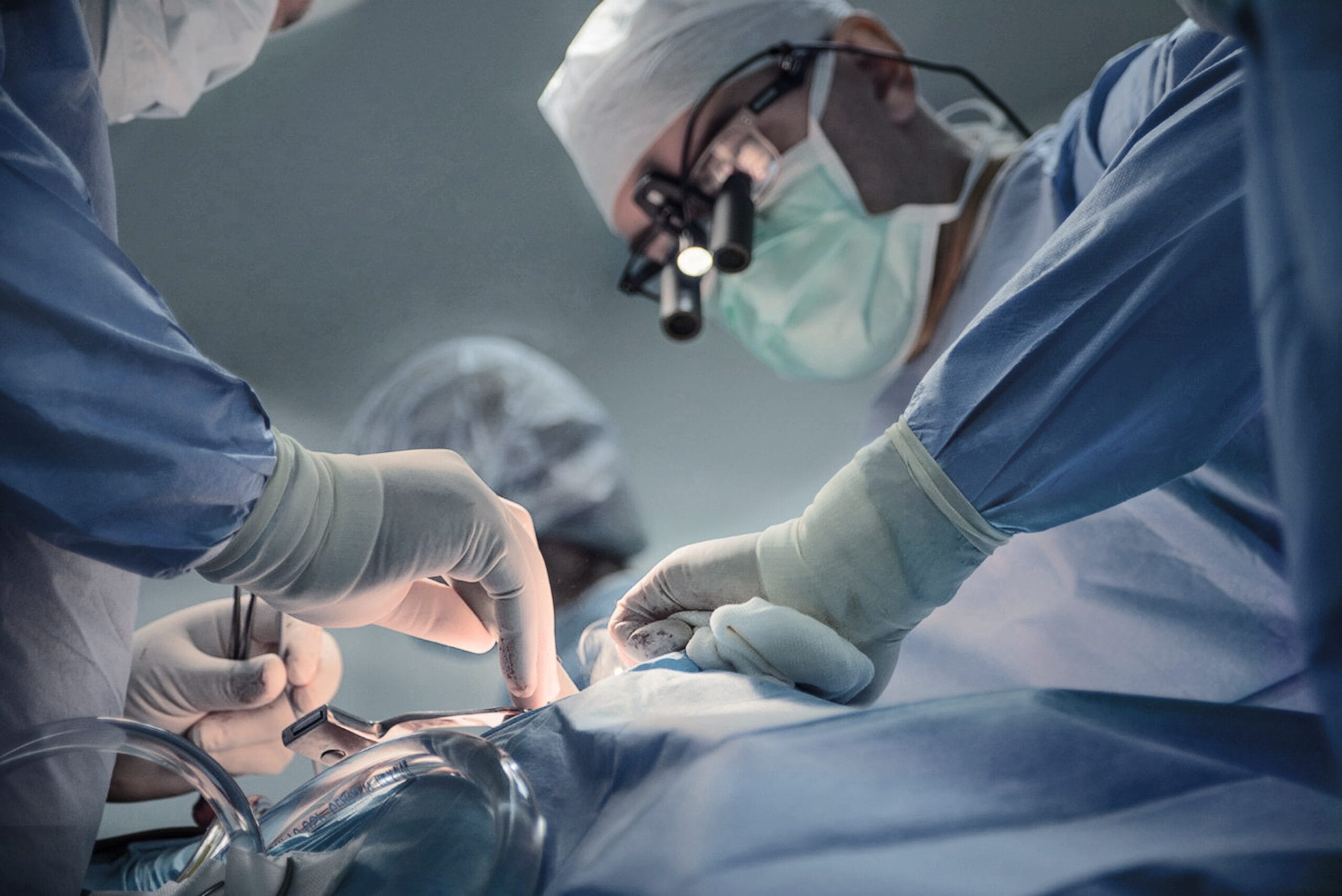 Coronary Artery Disease Singapore - A team of surgeons performing a surgical procedure in an operating room. The lead surgeon wears magnifying surgical loupes and a face mask, while assistants in sterile gloves carefully handle medical instruments.